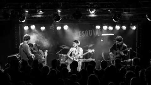 Bedford Esquires Black and white image of a pop group sitting in a smoky stage playing guitars