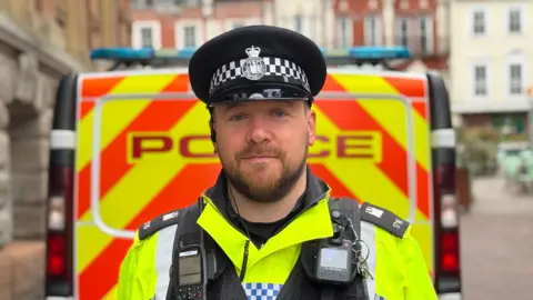 A close-up head and shoulders image of PC Matthew Dilks, who has a dark beard. He is wearing a police hat and uniform and standing in front of a police van. He is looking into the camera and smiling.