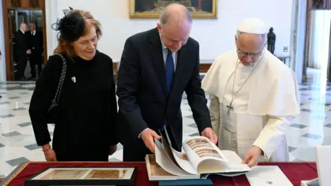 Vatican Media Micheál Martin, centre, standing beside Pope Leo and his wife Mary. They are standing behind a red table, flicking through a large book. 