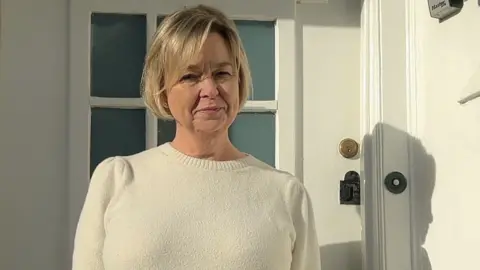 Lucy Green has short blonde hair. She wears a cream coloured jumper and stands in front of a door.