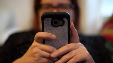 Closeup of the back of a mobile phone. A person is holding the phone with two hands. The person in the background is blurred out.