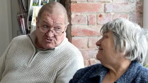 Alan Thompson is wearing a white jumper and has glasses with black edged rims. He is talking to Jane Innes who is wearing a blue jacket and is smiling back at him.