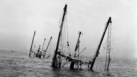 Getty Images The wreck of the SS Richard Montgomery is seen partially submerged in the Thames Estuary in an archive image from June 1962. Three large cargo masts and several smaller structures lean at different angles above the water, with rigging and cables hanging from them. The black-and-white photograph shows calm water stretching to the horizon beneath a pale sky.