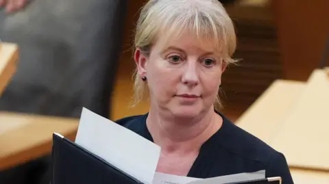 PA Media Shona Robison in the Holyrood chamber with a neutral expression on her face. She is holding a black leather folder with paper protruding from the top. She wears a navy top and has her blonde hair pinned up.