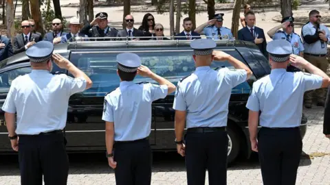 Getty Images A row of four police officers in uniform, seen from behind, salute a black hearse as it passes in front of them with flowers in the back