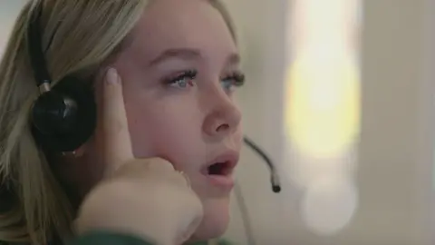 A call handler with a headset on has her hand against her face, with her mouth open, looking like she is in shock. She has blonde hair and blue eyes. 