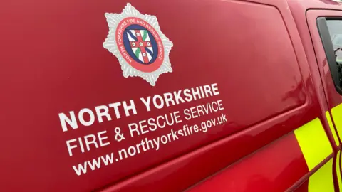 A red fire service vehicle with the crest of North Yorkshire Fire and Rescue Service and the service's name written on the van in white