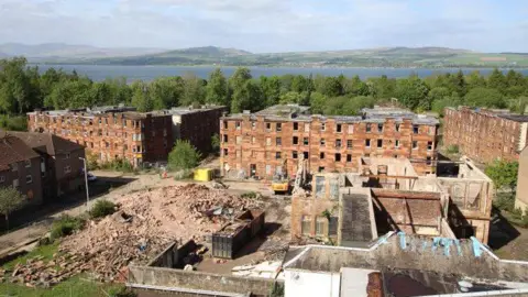 An aerial shot of a derelict red sandstone estate surrounded by trees with a river in the background - a number of the buildings are partly or fully demolished.