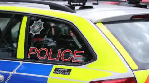Shows a yellow and blue police car with the word POLICE written on red on a side window below the PSNI crest.