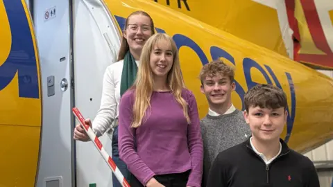 Four people, two women and two men, standing on steps leading out of an airplane. The group are all smiling at the camera. They are all different heights. The exterior of the plane is yellow. The door leading into the plane is open and exposes a white toilet door inside the plane.