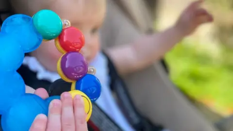 A toy in a baby's hand in the foreground, with the baby holding it out of focus. They are sitting in a pram with their other hand in the air. The toy is blue on one half and made up of green, red, purple, blue and yellow circles on the other half. The baby is dressed in a white romper. 