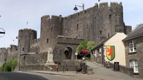 A tall stone castle with three visible towers and an arched entrance, next to a road.