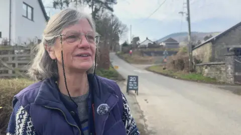 Fiona Cawley has a grey short bob which is half pulled back. She wears glasses attached to a spectacle cord and frowns into the camera while talking. 