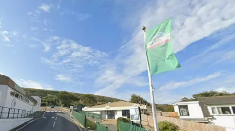 Google Google Street View of Alum Bay New Road outside the Needles car park and view point at Alum Bay. There is a flag pole by the side of the road flying a green flag bearing the Needles visitor attraction logo. There are three single-storey white buildings alongside the road which leads up a steep hill.