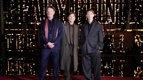 PA Media A man with a flat cap, blue suit and red patterned scarf stood next to a man with dark brown hair in a tweed coat and purple shirt and a man with brown hair and a dark grey suit. The double breasted jacket is buttoned up and the man has his hands in his pockets.