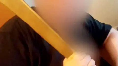 A heavily blurred photo of X, who is wearing a black T-shirt and holding a large machete