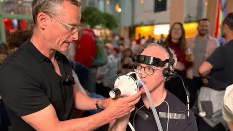 BBC Prof Damian Bailey holds a white breathing mask connected to clear tubing up to James' face, who is seated and wearing a head-mounted device with wires and sensors attached. James has additional medical-style electrodes taped to the chest and arm