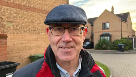 Martin Heath/BBC Stuart Copeland with glasses wearing a grey flat cap, grey shirt and red and grey anorak and smiling at the camera. He is standing in front of modern brick houses and a block-paved road