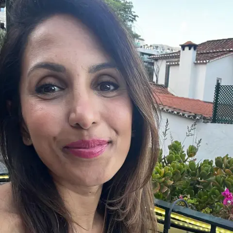 Handover A young woman sites outside on a sunny day - she has darker skin with brown hair, is waring some make up and is smiling at the camera 