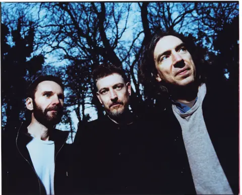 Tom Beard Promotional photo of Snow Patrol. The three members are standing against silhouetted tress, wearing a mixture of black and grey. 