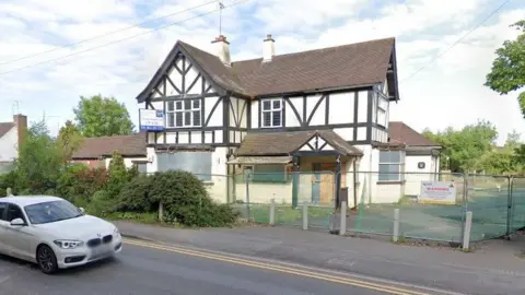 Google The shell of the fire-damaged pub, a mock-tudor black and white building dating back to the 1920s. Metal security fencing surrounds the site with a safety warning notice attached.