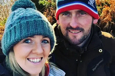 Ricky Rennie Ricky Rennie and wife Kirsten (left) are wearing woollen hats and jackets with autumn trees behind them. 