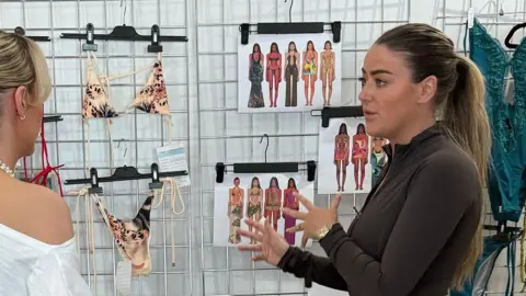 MURCI Olivia wearing a brown long sleeve top talking to another woman in front of a metal grid covered in bikinis and design sketches. 