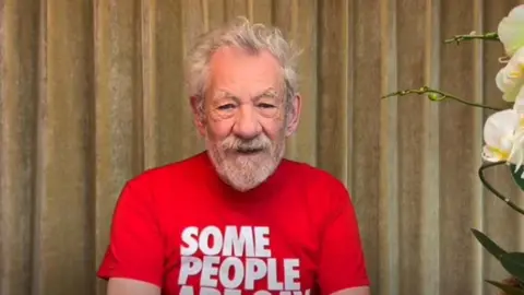 Screengrab of a video of Sir Ian McKellen sitting in front of a gold velvet curtain wearing a red tshirt. He has white hair an a neat beard
