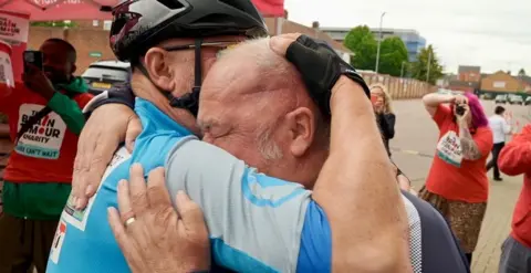 Mark Monaghan is on the left hugging one of his friends who just finished the bike ride. Mr Monaghan is wearing a navy blue T-shirt, and the other biker is wearing a light blue T-shirt with a black helmet.
