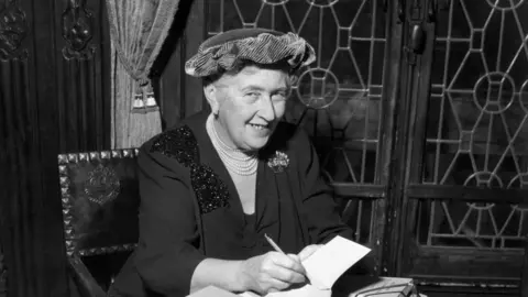 A black and white photo of Agatha Christie sitting on a chair as she signs a book. She is smiling at he camera and is wearing a hat, a black blouse and a pearl necklace.