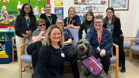 Nine women posing for a photo alongside a grey poodle with a purple coat on.