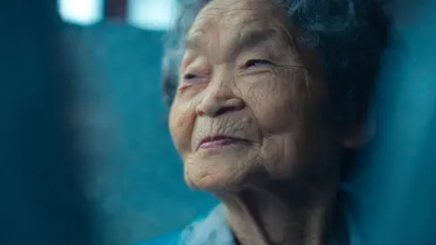 BBC/Minnow Films Portrait of Kiyomi Iguro, an elderly woman, looking to the left of the camera, with a small smile on her face. She has short greying hair. 