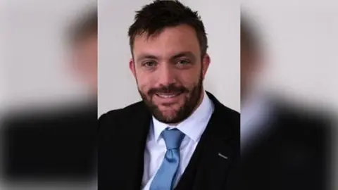 Pembrokeshire council Andrew Edwards looking at the camera and smiling. He has short cropped black hear, a trimmed dark beard and is wearing a dark suit and white shirt with a light blue tie