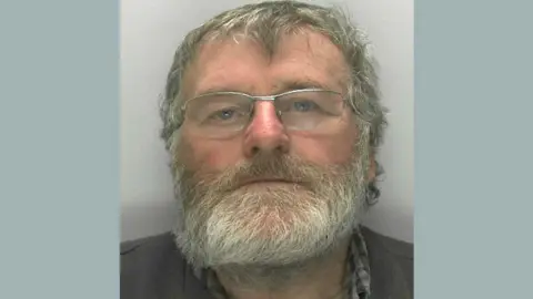 Gloucestershire Police Ramon Cleal looking at the camera for his custody mugshot. He has a white beard and is wearing glasses