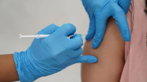 A person's hand- wearing blue rubber gloves - piercing a needle into another person's arm