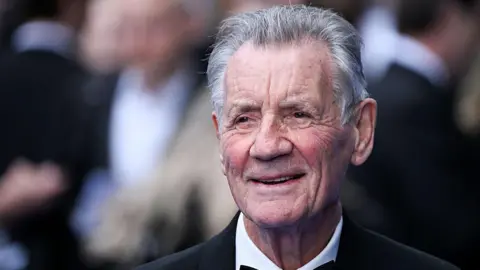 Michael Palin in a black suit with a bow tie against a blurred background