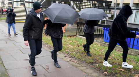 Image of Sophie Harvey and Elliot Benham outside Gloucester Crown Court. They are pictured under black umbrellas concealing their faces. 