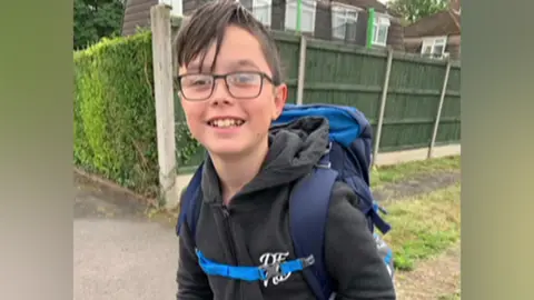 Family A boy in dark-framed glasses, in a dark hoodie, with a blue rucksack on his ban. In the background there is a hedge and fencing surrounding a house