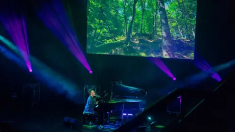 The Fire Station Emeli Sandé playing the piano and singing on a purple-lit stage with a forest projected on a screen above her. She is wearing a black shirt and her hair is up. 