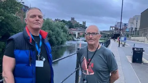 BBC Toby Jones, left, and Gav are two middle-aged men. Toby is wearing a dark blue gilet and black t-shirt and Gav has spectacles and is wearing a grey t-shirt. Both have close-cropped hair and are wearing lanyards with an identity badge. They are standing on a street next to a railing with the river to one. Durham Cathedral and Castle can be seen in the far background.