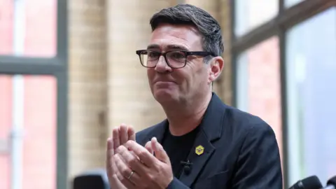 Andy Burnham clapping at a speech he gave in Manchester. He is wearing a dark t-shirt and jacket, with a yellow badge pinned to his lapel.