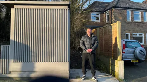 Brett Kemp, a man with short grey hair wearing a dark puffer jacket, standing next to the metal shed-style structure with corrugated sides. His house can be seen in the background