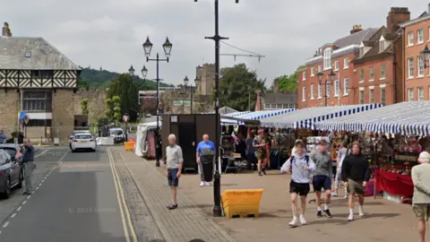 A road running alongside a paved area with people walking around and market stalls with blue and white striped awnings and a castle and a black and white timbered building in the background