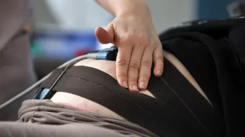 A generic close-up of a female medic feeling a pregnant patient's womb, which is belted with dark straps for an examination.
