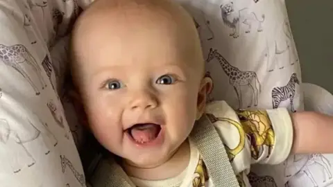 A baby sitting in a white baby seat with animals on it is smiling at the camera