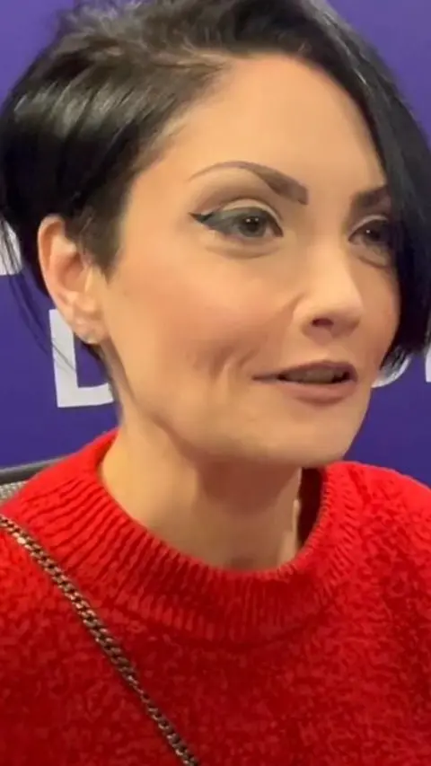 Gillian Nair looking to the right. She has short black hair and is wearing a red jumper.