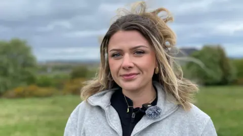 A young woman stands outdoors in a grassy area, facing the camera, with wind‑blown hair visible against a cloudy sky. Trees and distant buildings appear softly blurred in the background, while the person wears a light grey fleecy zipper top.