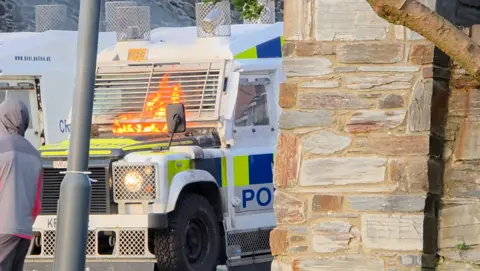 Shows a white police Land Rover with blue and yellow squares on the side and a youth in a grey tracksuit to the left