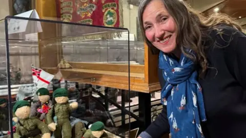 BBC Tansy Forster, wearing a blue scarf and navy jumper, pictured next to knitted British soldiers depicting the D-Day landings in Normandy, France, 1944. The soldiers are dressed in light green uniforms and dark green berets, while their faces and hands are cut. white. The knitted soldiers are also carrying the Union Jack/St George's Cross flag.