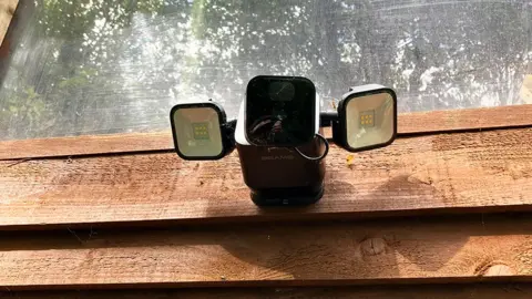 BBC A black security camera with two lights attached to the side of a brown shed
